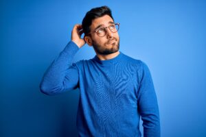 Young handsome man with beard wearing casual sweater and glasses over blue background confuse and wondering about question Uncertain with doubt, thinking with hand on head Pensive concept