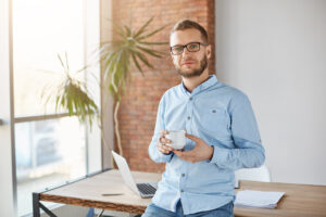 Close up portrait of young attractive company founder in glasses and casual outfit, standing in personal office, holding cup of coffee in hands, looking in camera with relaxed expression, posing for company website photo shoot